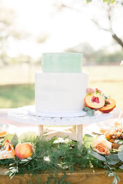 Picture of a two tier cake. A short 6 inch round cake with pastel mint smooth and sharp edged buttercream. On top of an 8 inch round cake with white buttercream decorated smoothly with sharp edges. A halved peach as well as florals and greenery act as decor on the right side