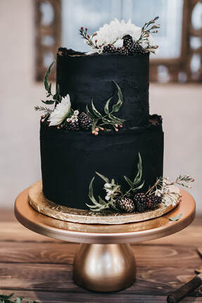 Picture - A 6 inch round and 8 inch round two tier cake. With black stucco textured buttercream, florals, greenery, and gold dusted blackberries. All placed on a round gold metal 12
