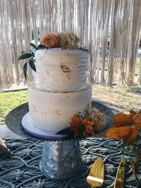 Picture of a 6 inch round with fat horizontal buttercream ruffles on an 8 inch with smooth buttercream and rough edges. Both are decorated with white pearl sprinkles, edible gold leaf, and fall themes flowers and greenery.
