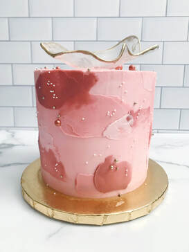 Picture of a 6 inch cake with a monochromatic pink color scheme. Small white sprinkles, gold and pink round sprinkles, and a isomalt sail with gold edges on top.