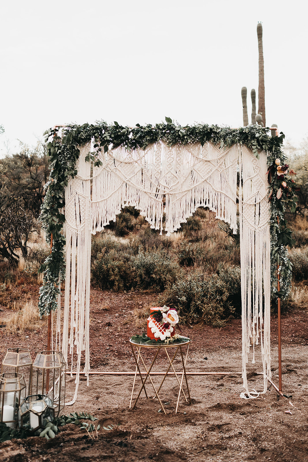 Picture of dessert wedding. A copper arch draped with macrame and greenery. Underneath is a rusty orange to tier cake made of a small 6 inch cake and an 8 inch cake. Decorated with piped buttercream macrame and florals. Glass lanterns placed on either side. 