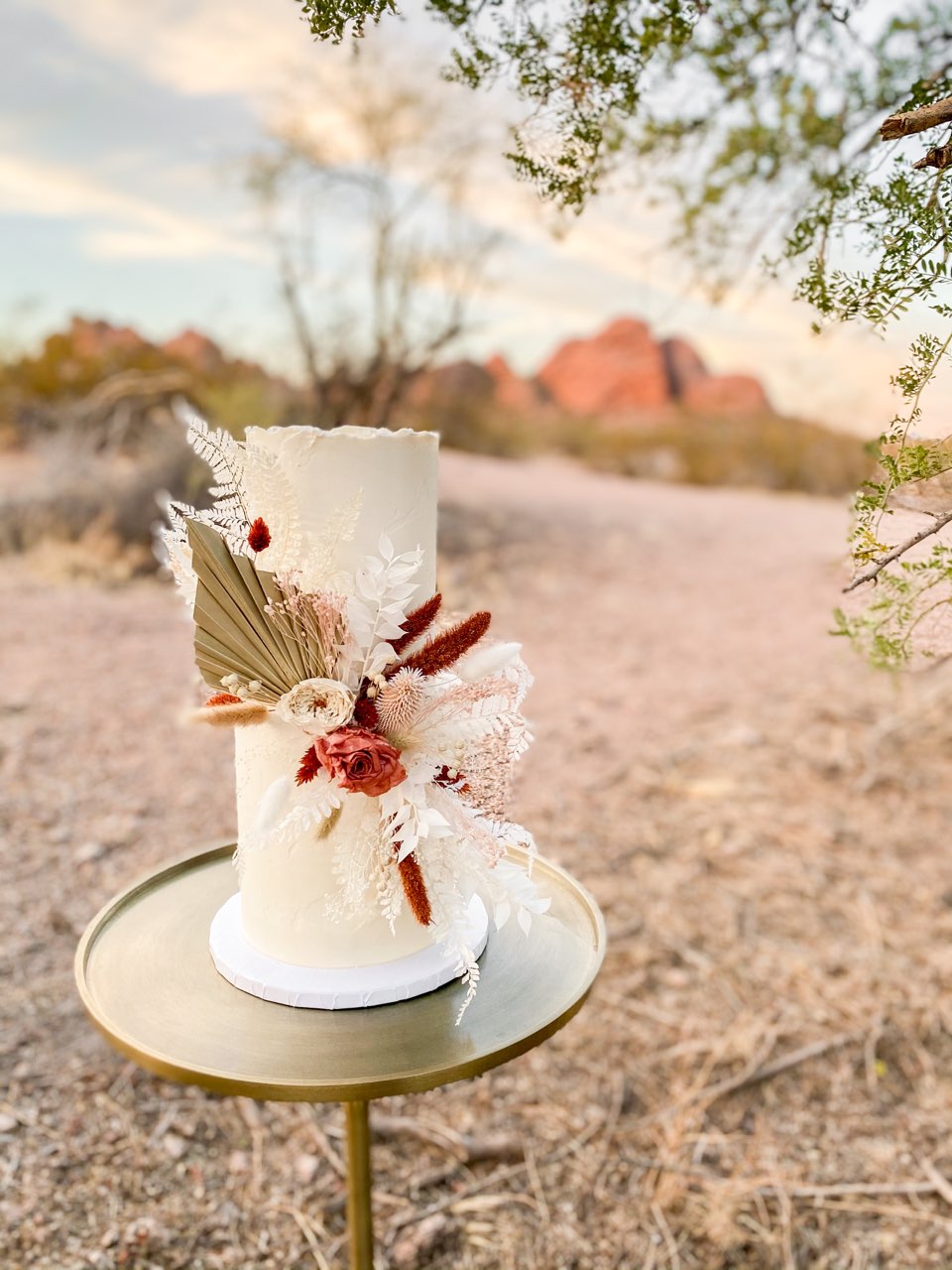 Picture of a tall 5 in and tall 6 inch tiered cake with textured buttercream and dried floral accesories. pictured in the desert ontop of a gold cake stand