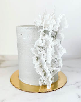 Picture of a tall 6 inch cake with concrete colored and textured buttercream and sharp edges. Decorated with vertical ascending fluffy rice paper sails.