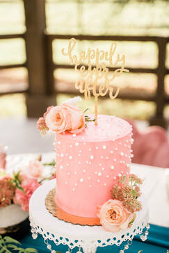 Picture of a tall 6 inch cake smooth pink buttercream and sharp edges. Decorated with pink flowers, multiple sizes of pearl sprinkles, and a gold glitter topper that reads 