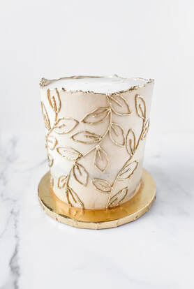 Picture - A tall 6 inch cake with smooth sides and rough edge buttercream top. Decorated with piped trailing leaf design painted gold.