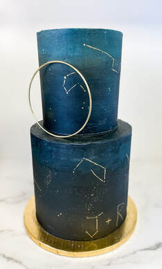 Picture of a tall 6 inch and tall 8 inch cake with a night sky ombre and hain painted constellations. The bride and grooms initials are also drawn as constellations. the cake is accented with a gold hoop placed in the bottom tier.